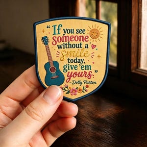 May include: A shield-shaped embroidered patch with a yellow background and dark blue border. It features a teal acoustic guitar, a sun icon, small hearts, and floral accents. The text reads: If you see someone without a smile today, give 'em yours. - Dolly Parton.