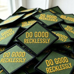 May include: A collection of diamond-shaped olive green fabric patches with black stitched borders. Each patch features the text DO GOOD RECKLESSLY embroidered in bold yellow capital letters, centered across two lines on the textured surface.