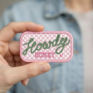 May include: A rectangular embroidered patch with rounded corners featuring a white and light pink checkered background. The text "Howdy" is stitched in green cursive script above the pink block-lettered word "HONEY" and a small pink heart.