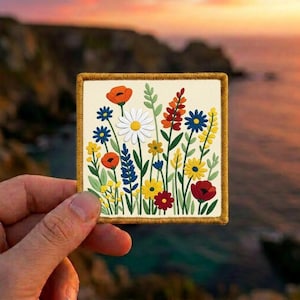 May include: A square embroidered patch featuring a cream-colored background with a vibrant botanical illustration of various wildflowers including red poppies, white daisies, blue cornflowers, and yellow blossoms with green stems and leaves.