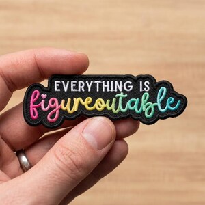 May include: A black embroidered patch with a scalloped border featuring the white text EVERYTHING IS and the word figureoutable in a gradient of pink, yellow, green, and blue cursive script with a small heart above the letter i.