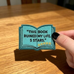 May include: A teal embroidered patch shaped like an open book with black text that reads, "THIS BOOK RUINED MY LIFE. 5 STARS." and five yellow embroidered stars underneath the text. The patch features a dark teal border and detailed stitching.