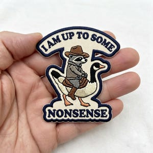 May include: An embroidered patch featuring a raccoon wearing a brown cowboy hat and outfit, riding a white and brown goose. The patch has a cream-colored background with a dark blue border and the text I AM UP TO SOME NONSENSE in dark blue.