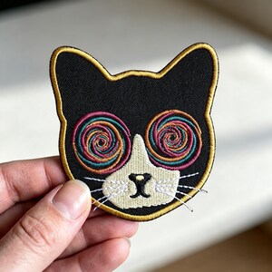 May include: An embroidered cat-shaped patch featuring a black head with a gold border. The cat has a white muzzle, a black nose, and white whiskers. The eyes are depicted as large, colorful spirals in shades of pink, orange, and teal, creating a hypnotic effect.