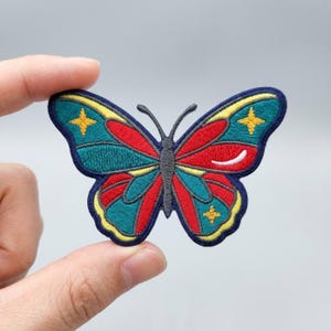 May include: An embroidered butterfly patch featuring a dark blue border, teal wings with red accents, and yellow four-pointed star details. The central body is dark gray, and a small white crescent shape is visible on the right wing section.