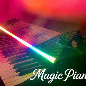 Op de afbeelding: Een zwart elektronisch keyboard met een lange, gloeiende LED-lichtbalk over het bovenpaneel die overgaat van oranje naar groen. Het keyboard heeft een bedieningspaneel met knoppen en een scherm, en de tekst Magic Piano staat in het wit geschreven.