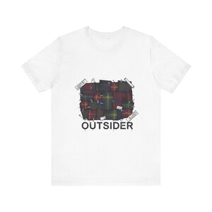 Può includere: T-shirt bianca con un motivo a quadri, spille di sicurezza e nastro adesivo, e la parola "OUTSIDER" in nero. Il motivo a quadri comprende tonalità di rosso, verde e giallo.
