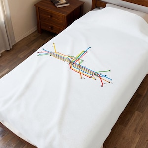 Op de afbeelding: Wit sprei met een kleurrijk metrokaartontwerp. De kaart gebruikt een netwerk van lijnen in verschillende kleuren, waaronder rood, blauw, geel en groen, om routes weer te geven. De sprei ligt op een houten bedframe.