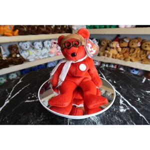 Puede incluir: Un oso de peluche rojo con gorro de aviador marrón con gafas, bufanda blanca y emblema circular. El oso tiene una etiqueta en forma de corazón Ty. El oso está sentado en un espejo redondo.