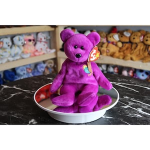 Puede incluir: Un oso de peluche morado con una nariz negra y una etiqueta. El oso está sentado en un plato blanco y rojo. Otros peluches son visibles en el fondo en estantes.