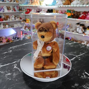 Puede incluir: Un oso de peluche marrón con un gorro de aviador marrón con gafas, una bufanda blanca y una chaqueta marrón. El oso está dentro de una vitrina de plástico transparente. Una etiqueta roja está adherida al oso. El oso tiene un parche que dice "The World of Ty".