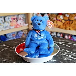 Könnte beinhalten: Ein blauer Plüschbär mit schwarzer Nase und Augen, der ein Band mit dem Wort "NASCAR" trägt. Der Bär sitzt auf einer weißen, kreisförmigen Plattform. Ein rotes und weißes Etikett ist am Ohr des Bären befestigt. Weitere Plüschtiere sind im Hintergrund zu sehen.