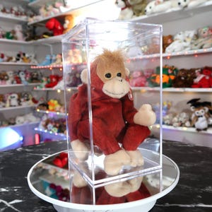 以下が含まれることがあります： 赤い体、ベージュの顔、タグが付いたぬいぐるみオランウータン。透明なアクリルボックスの中に展示されています。おもちゃは回転する鏡面の上にあります。背景には、他のぬいぐるみでいっぱいの棚が見えます。