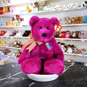 Puede incluir: Un oso de peluche magenta con un lazo dorado y una etiqueta Ty. El oso tiene una nariz negra y un parche bordado. Está sobre una superficie blanca, con otros peluches en el fondo. Un objeto de colección.