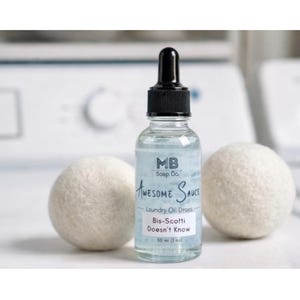 Puede incluir: Una botella de vidrio transparente de gotas de aceite para lavandería con tapón cuentagotas negro, etiquetada como "Awesome Sauce" y "Bis-Scotti Doesn't Know". Dos bolas de secado de lana blanca flanquean la botella. Contiene 30 ml.