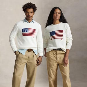 May include: Two white long-sleeved crewneck sweaters featuring a red, white, and blue American flag graphic on the chest with the letters RL embroidered in red. The sweaters are paired with light blue collared shirts and khaki trousers.