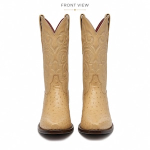 May include: A pair of tan cowboy boots featuring a textured quill pattern on the foot and intricate tonal stitching on the tall shaft. The boots have a pointed toe, a dark brown sole, and a deep red interior lining. The text FRONT VIEW is visible.