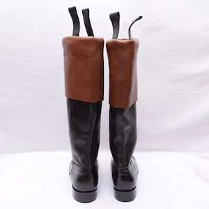 May include: A pair of tall boots featuring black leather lower sections and brown leather fold-over cuffs at the top. Each boot includes two black pull tabs extending upward from the rear of the collar, with a smooth finish on the black leather and a textured grain on the brown leather cuffs.