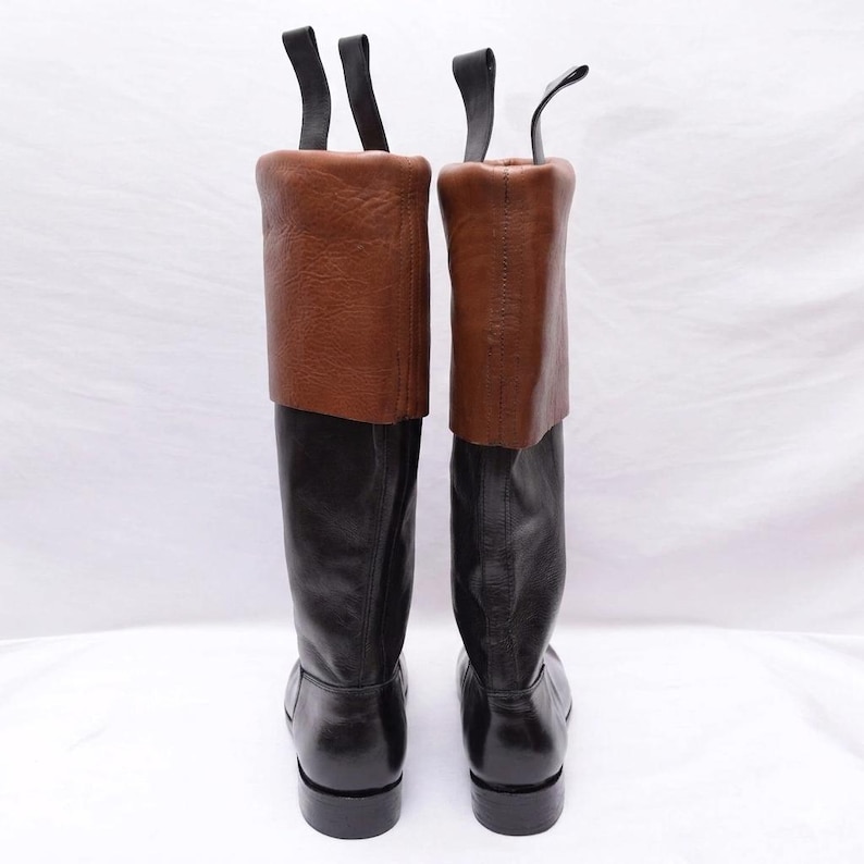 May include: A pair of tall boots featuring black leather lower sections and brown leather fold-over cuffs at the top. Each boot includes two black pull tabs extending upward from the rear of the collar, with a smooth finish on the black leather and a textured grain on the brown leather cuffs.