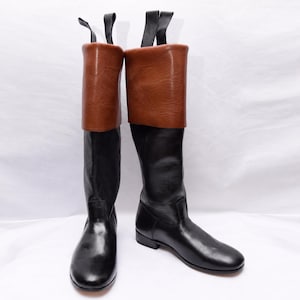 May include: A pair of knee-high boots featuring a smooth black lower section and a contrasting tan brown fold-over cuff at the top. Each boot includes a black pull tab extending upward from the rear collar, with a rounded toe and low heel.