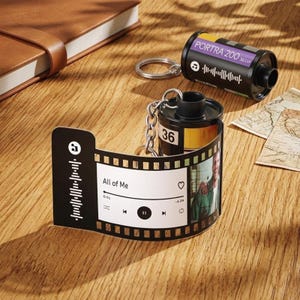 May include: A black film roll keychain and a film strip-shaped photo display. The film roll is labeled "PORTRA 200" and has a music-themed graphic. The photo display shows a photo and the text "All of Me".