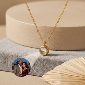 Puede incluir: Un collar dorado con un colgante redondo. El colgante tiene un centro claro y circular. El collar se exhibe sobre una superficie de tonos neutros. Una pequeña foto circular de una pareja está en la esquina inferior izquierda.