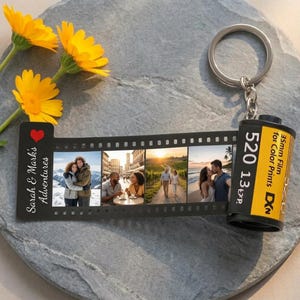 Puede incluir: Un llavero personalizado con un diseño de tira de película que muestra cuatro fotos. La tira de película negra tiene el texto "Sarah & Mark's Adventures" y un corazón rojo. El llavero incluye un cartucho de película de 35 mm con el texto "520 13 exp".