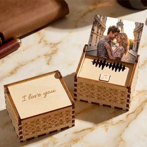 May include: Two wooden music boxes. One box is closed with the words "I love you" engraved on the lid. The other box is open, displaying a photo of a couple and a sound wave cutout. The boxes have decorative patterns.
