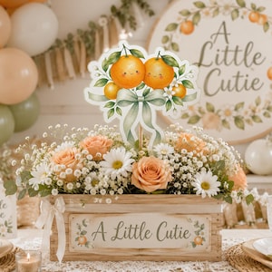 Könnte beinhalten: Holzkiste mit Blumenschmuck und dem Text "A Little Cutie". Die Anordnung umfasst Pfirsichrosen, Gänseblümchen und Schleierkraut. Ein Schild mit dem gleichen Text und Orangen-Design ist im Hintergrund. Pfirsich-, weiße und grüne Ballons.