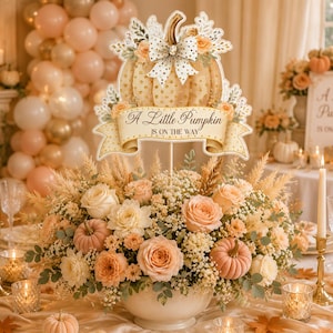 Könnte beinhalten: Ein dekoratives Herzstück mit einem kürbisförmigen Schild mit dem Text A Little Pumpkin IS ON THE WAY, verziert mit einer weiß gepunkteten Schleife, eingebettet in ein Blumenarrangement aus pfirsichfarbenen Rosen, weißen Blüten und kleinen Kürbissen.