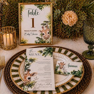 May include: A baby shower table setting with a giraffe theme. Features a framed table number, menu, place card, and giraffe figurine. Gold cutlery, a candle holder, and glassware complete the elegant arrangement.