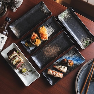 Puede incluir: Una colección de platos rectangulares para sushi en negro, marrón y blanco, con diseños moteados y pintados. Los platos están colocados sobre una superficie de madera, algunos con rollos de sushi y guarniciones. Se ve una botella de sake japonés.