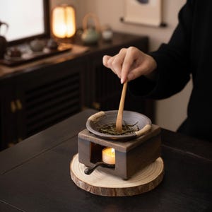 Pode incluir: Um aquecedor de chá de madeira escura com uma pequena tigela de cerâmica contendo folhas de chá. Uma vela acesa está dentro do aquecedor, e uma colher de madeira mexe o chá. O aquecedor repousa sobre uma fatia de madeira.