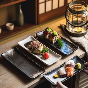 Puede incluir: Una colección de platos de sushi rectangulares de cerámica en varios colores y texturas. Los platos están llenos de sushi, sashimi y guarniciones. Los platos están dispuestos sobre una superficie de madera, creando una presentación visualmente atractiva.