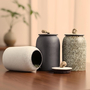 Puede incluir: Una colección de recipientes de cerámica en tonos neutros. El conjunto incluye una pieza cilíndrica beige moteada con una abertura, un tarro negro mate y un tarro verde-gris moteado, todos con tapas atadas con cuerda. Los artículos están dispuestos sobre una superficie de madera.