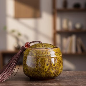 May include: A small, round ceramic jar with a lid, featuring a speckled yellow and brown glaze. A brown tassel with a red bead hangs from the lid. The jar is likely for storing small items or as a decorative piece.