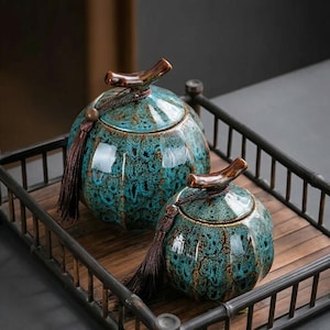 Peut inclure: Deux pots en céramique avec couvercles, présentant un glaçage moucheté bleu-vert et marron. Chaque pot a une poignée en forme de tige marron et un gland assorti. Les pots sont présentés sur un plateau en bois avec un cadre en métal foncé.