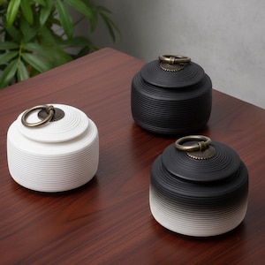 Pode incluir: Três potes de cerâmica com tampas. Um é branco e dois são pretos com um gradiente para branco na base. Cada pote tem uma alça circular de latão na tampa. Os potes estão em uma superfície de madeira escura.