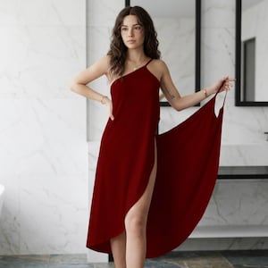 May include: A deep red, sleeveless, one-shoulder dress featuring a high side slit that extends up the thigh. The garment has a fluid, draped silhouette with a single thin strap over the shoulder, creating an asymmetrical neckline and hemline.