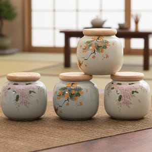 Puede incluir: Cuatro tarros de cerámica con tapas de madera apilados sobre una superficie marrón. Los tarros son de color verde claro con diseños florales en naranja y rosa. Los tarros son redondos con un acabado liso.