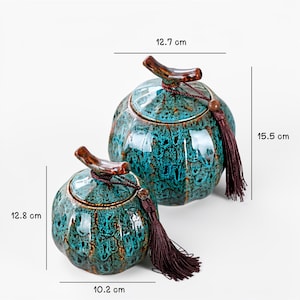May include: Two ceramic jars with lids, featuring a teal and brown mottled glaze. The larger jar is 15.5 cm tall and 12.7 cm wide, while the smaller jar is 12.8 cm tall and 10.2 cm wide. Each jar has a brown wooden handle and a tassel.