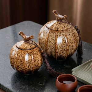 May include: Two ceramic jars with lids. The jars are round with a speckled brown glaze and vertical brown stripes. Each jar has a curved handle on the lid and a decorative tassel. The jars are on a dark surface.