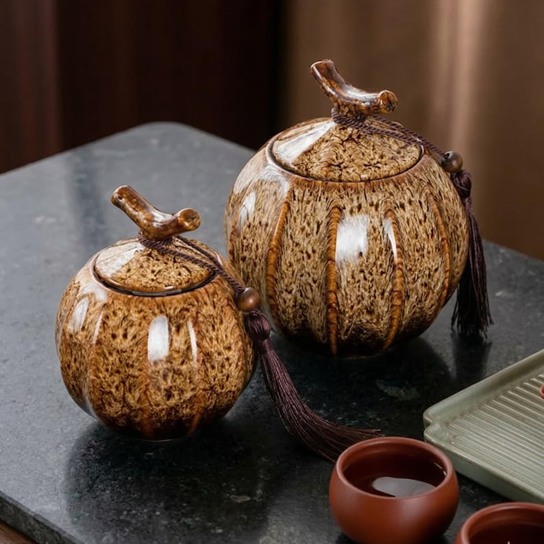 May include: Two ceramic jars with lids. The jars are round with a speckled brown glaze and vertical brown stripes. Each jar has a curved handle on the lid and a decorative tassel. The jars are on a dark surface.