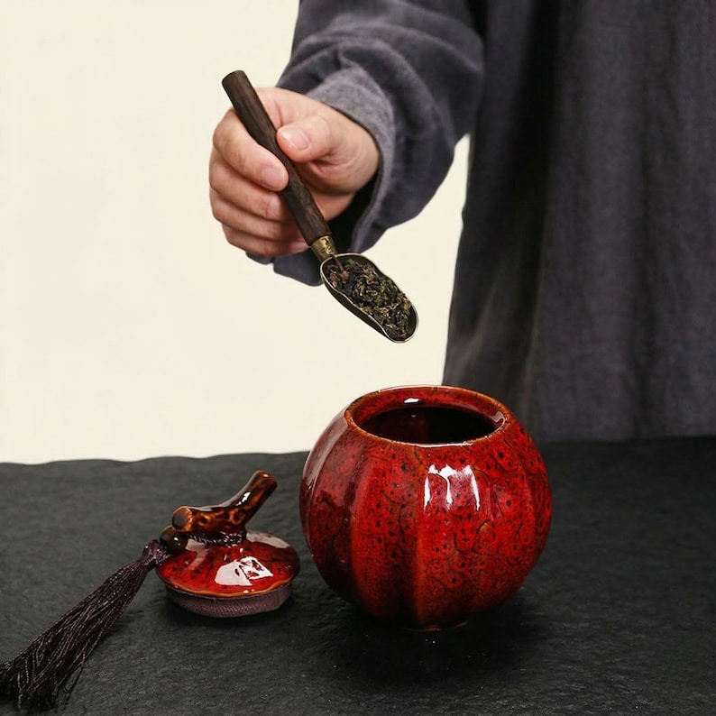 May include: A red ceramic tea caddy with a matching lid and a dark tassel. A wooden and metal scoop holds loose tea leaves above the caddy. The caddy has a textured surface and a round shape.