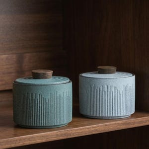 Op de afbeelding: Twee keramische potten met deksels op een houten plank. De potten zijn cilindrisch met verticale getextureerde lijnen en een teal en grijze kleur. Elke pot heeft een houten knop bovenop het deksel. De achtergrond is een donkere houten kast.