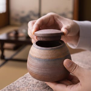 Puede incluir: Un tarro de cerámica marrón hecho a mano con tapa. El tarro tiene forma redondeada con líneas horizontales y una banda de color azul grisáceo. La tapa es de madera. El tarro está siendo sostenido por dos manos.