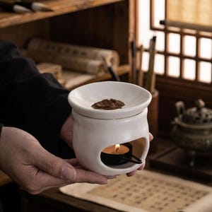 Könnte beinhalten: Ein weißer Keramik-Diffusor für ätherische Öle mit einer kleinen Kerze im Inneren. Der Diffusor hat eine runde Oberseite mit einem kleinen Haufen braunen Materials. Der Diffusor wird in den Händen einer Person gehalten.