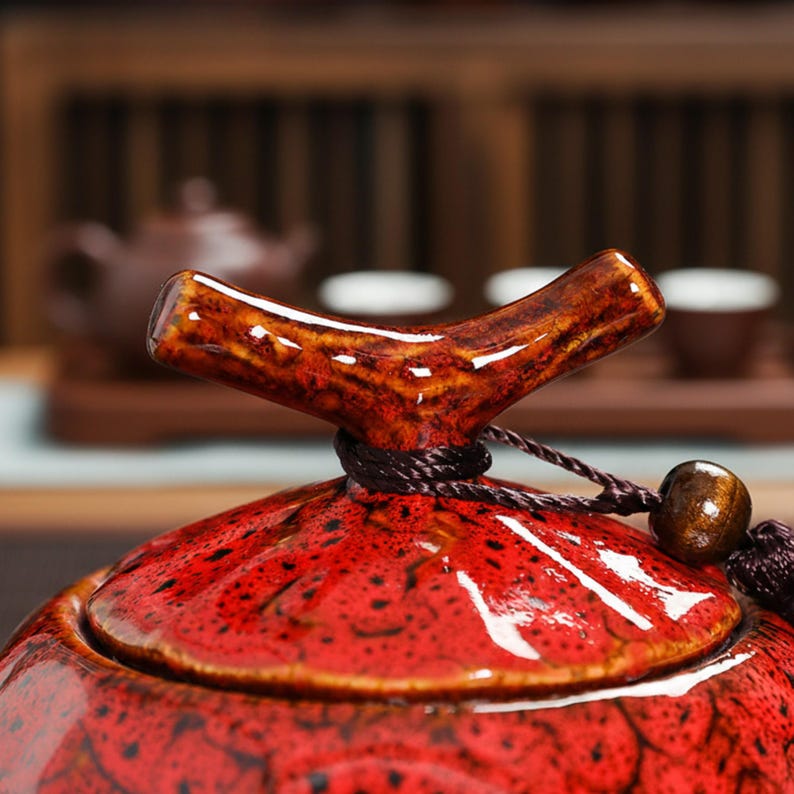May include: A close-up of a red ceramic tea caddy with a textured surface and a brown, branch-shaped handle. The lid is secured with a dark brown cord and a wooden bead. The background shows a tea set.