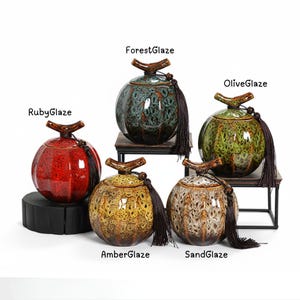 May include: Five ceramic jars with lids and tassels. The jars are in shades of red, green, yellow, and brown. Each jar has a unique glaze, with the names "RubyGlaze", "ForestGlaze", "OliveGlaze", "AmberGlaze", and "SandGlaze" written above them.