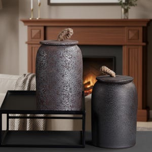 May include: Two ceramic jars with lids, one dark reddish-brown and the other black. Both have a textured finish and rope handles on the lids. They sit on a black metal tray. The jars are suitable for home decor.